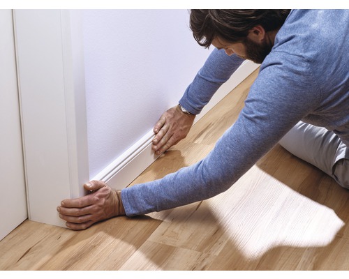 Een man installeert een witte plint tegen een muur in een kamer met een houten vloer.