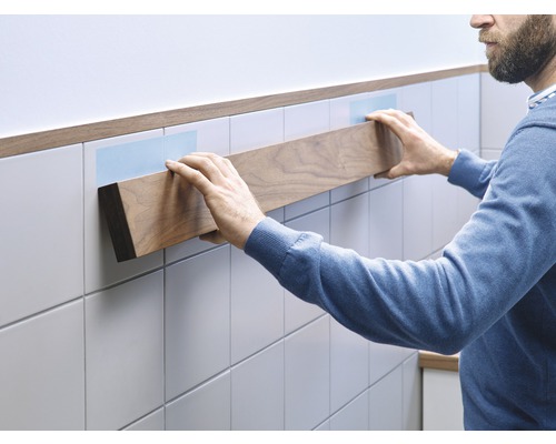 Man bevestigt een houten plank aan een betegelde muur met plakstrips.