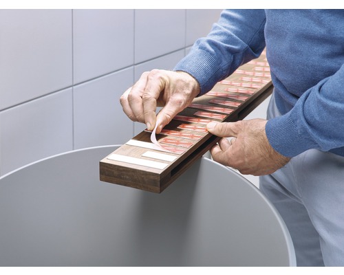 Een persoon verwijdert de beschermfolie van een plakstrip op een houten lat boven een afvalemmer.