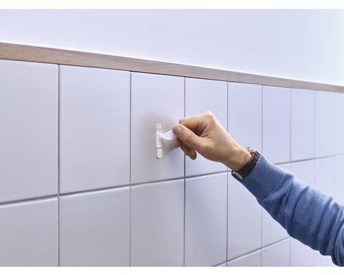 Iemand bevestigt een zelfklevende haak aan een betegelde wand.