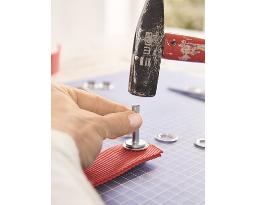 Iemand bevestigt met een hamer een oogje aan een stuk stof.