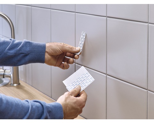 Een persoon bevestigt dubbelzijdige plakstrips op een betegelde muur.