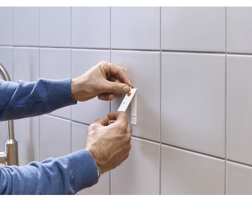 Een persoon bevestigt een voorwerp met een plakstrip aan een betegelde muur.