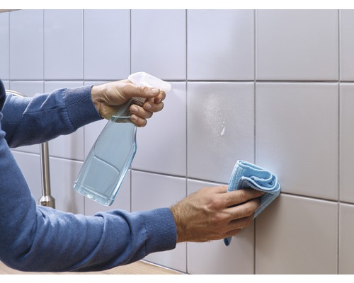 Een persoon reinigt tegels met een reinigingsspray en een doek.