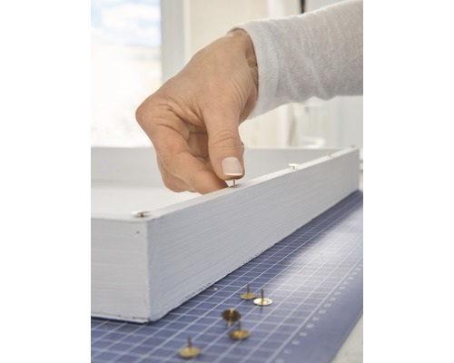 Een hand bevestigt een punaise aan een wit houten frame.