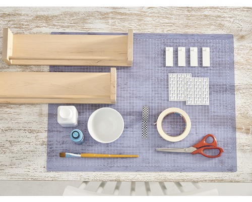 Twee houten planken, verf, penseel, schaar, tape en decoratief papier liggen op een snijmat.