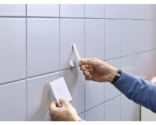 Een man bevestigt een voorwerp aan een betegelde wand met dubbelzijdige tape.