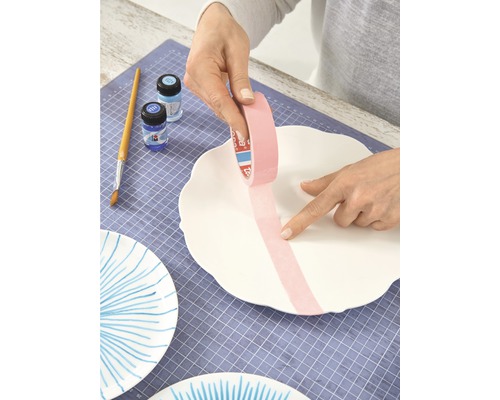 Een persoon plakt schilderstape op een witte keramische plaat om deze te decoreren. Penseel, kleuren en meer beschilderde platen zijn zichtbaar.