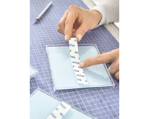Persoon bevestigt een Tesa plakstrip op een transparant oppervlak op een knutselmat.