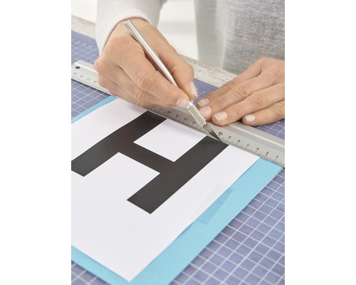 Iemand snijdt een letter uit papier met een hobbymes en een liniaal.