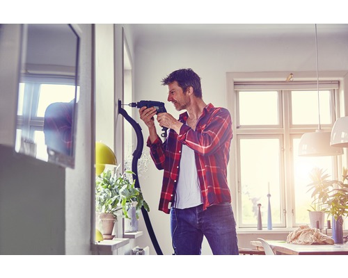 Man boort met een boormachine een gat in de muur, terwijl een stofzuiger het stof opzuigt.