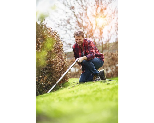 Een man knielt in de tuin en snoeit een haag met een heggenschaar.