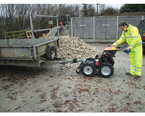Persoon bedient een minitransporter die een aanhanger trekt. Op de achtergrond ligt een stapel stenen.