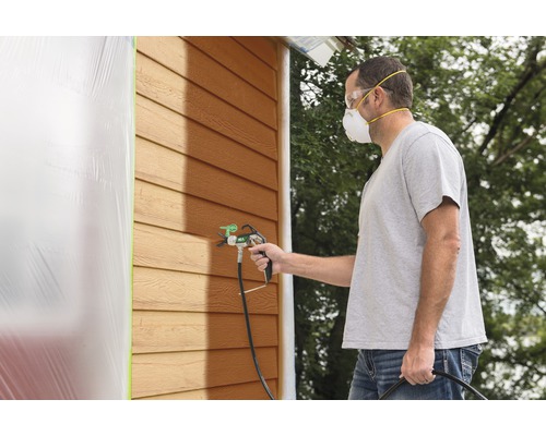 Man schildert huisgevel met verfspuit en draagt beschermend masker en bril.
