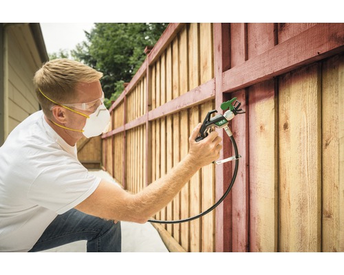 Man schildert houten schutting met verfspuit, met veiligheidsbril en ademhalingsmasker.