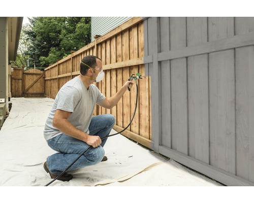 Een persoon schildert een houten schutting met een verfspuitpistool.