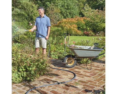 Een man besproeit planten met een tuinslang naast een kruiwagen met tuingereedschap.