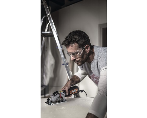 Een man met een veiligheidsbril zaagt een werkstuk met een cirkelzaag. Op de achtergrond staat een ladder.