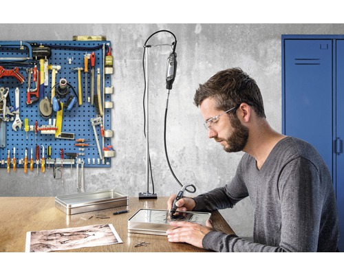 Man graveert met een Dremel gereedschap in een metalen doos op een bureau, gereedschapswand op de achtergrond