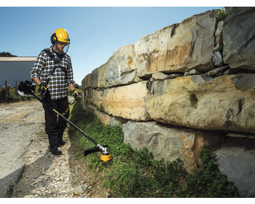 Man gebruikt een bosmaaier om gras langs een stenen muur te trimmen.