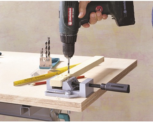 Een persoon boort met een accuboormachine in een stuk hout dat met een bankschroef is vastgezet.