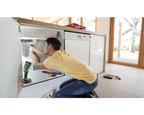 Een man repareert een afvoer onder een gootsteen met gereedschap en een werklamp in een lichte keuken.