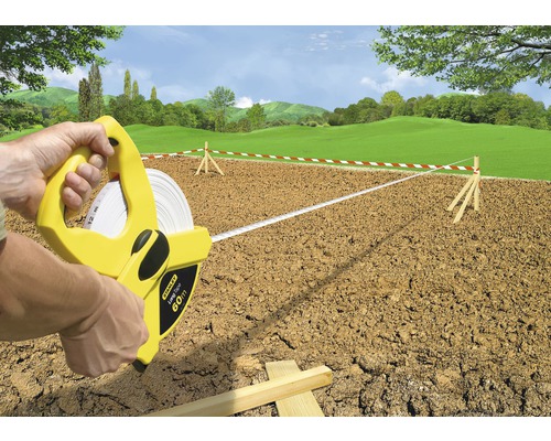 Een persoon gebruikt een 60 meter lange rolmaat van het merk Stanley om een terrein te meten dat is gemarkeerd met rood-witte linten.