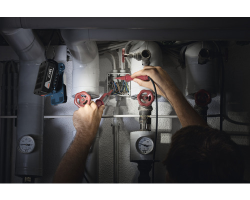 Een installateur werkt met spanningstesters aan een verdeelkast, verlicht door een bouwlamp.