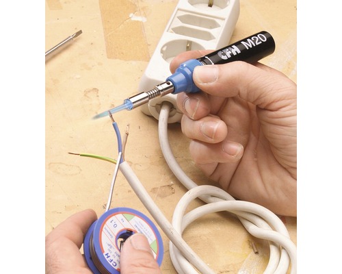 Een hand soldeert een elektrische leiding met een soldeerbout. Op tafel liggen een spoel soldeertin en een stekkerdoos.