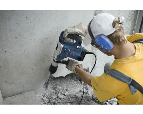 Een man werkt met een breekhamer op een betonnen muur. Hij draagt beschermende handschoenen en gehoorbescherming.
