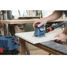 Een persoon schuurt een houten plank met een Bosch vlakschuurmachine, aangesloten op een stofzuiger.