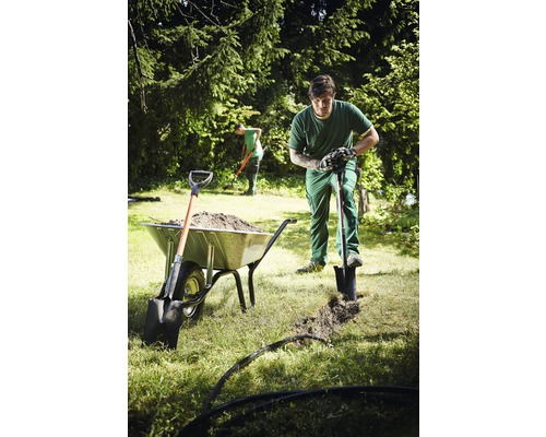 Tuinman graaft een geul met een schop naast een kruiwagen vol aarde.