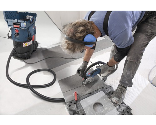 Een vakman snijdt een tegel met een haakse slijper, waarbij een industriële stofzuiger stof afzuigt.