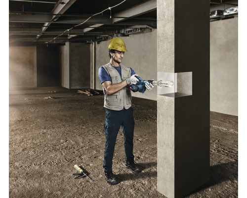 Een man met een veiligheidshelm boort met een boorhamer in een betonnen pilaar op een bouwplaats.