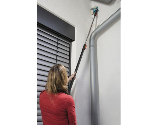 Een vrouw gebruikt een telescopische borstel om een gevel naast een regenpijp schoon te maken.