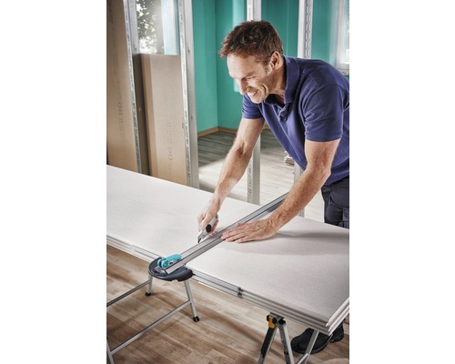 Een man snijdt een gipsplaat met een mes en een liniaal op een tafel.