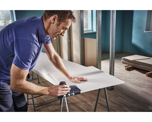 Man bewerkt gipsplaat met een kantenschaaf op een werkbank in een ruwbouw.