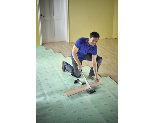 Een man installeert laminaat met een trekijzer en contactgeluidsisolatie in een kamer.