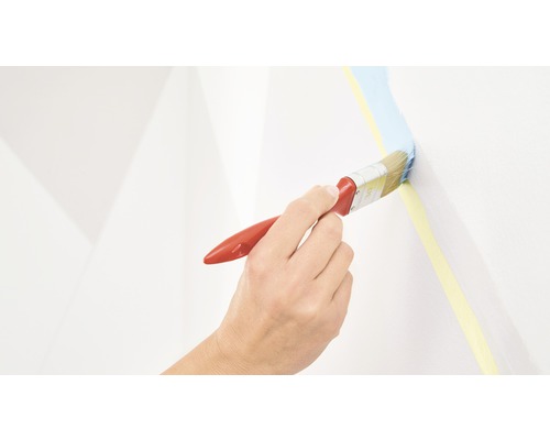 Een hand schildert met een kwast blauwe verf langs een gele schilderstape op een muur.