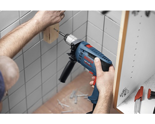 Ambachtsman boort met een klopboormachine een stuk hout aan een muur. Op de achtergrond is een kast te zien. Op de grond liggen schroeven.