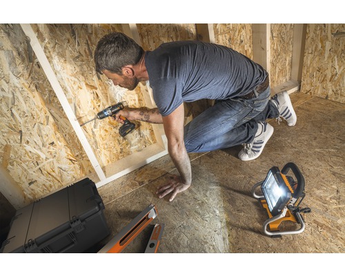 Ambachtsman werkt met een accuschroefboormachine in een houten casco, verlicht door een bouwlamp.