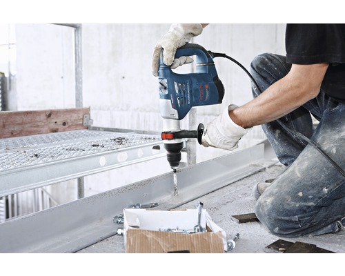 Een man boort met een boormachine in een metalen stuk. Schroeven en pluggen liggen klaar.