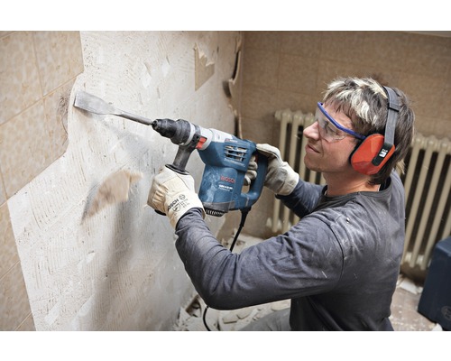 Een man gebruikt een sloophamer om tegels van een muur te verwijderen. Hij draagt een veiligheidsbril en gehoorbescherming.