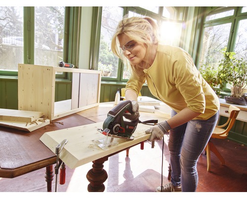 Vrouw zaagt houten plank met een decoupeerzaag op een houten tafel. Op de achtergrond staat een onafgewerkte houten plank.