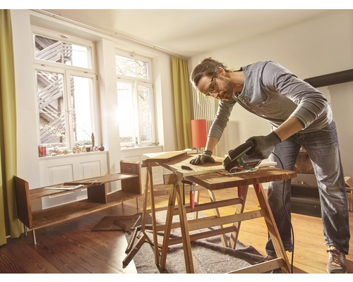Een man werkt met een multifunctioneel gereedschap op een houten oppervlak in een ruimte met planken en ramen.