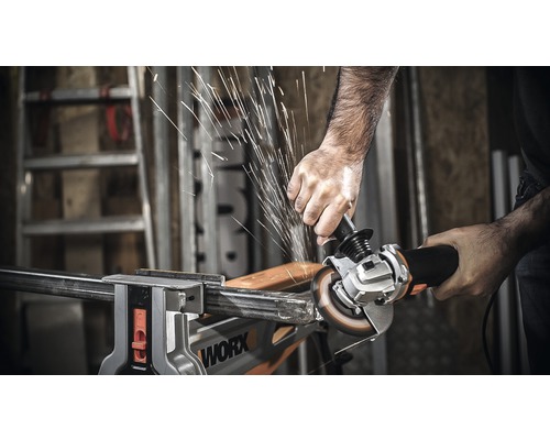 Iemand snijdt een stuk metaal met een haakse slijper, dat in een bankschroef is vastgeklemd. Er komen vonken vrij.