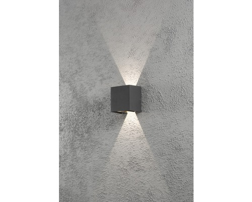 Kubusvormige wandlamp straalt licht naar boven en beneden op een muur.