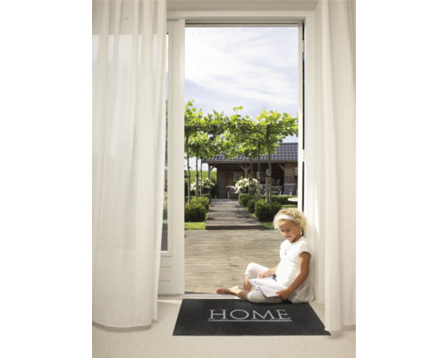 Een jong meisje zit te lezen op een deurmat met het woord ''Home'' voor een open terrasdeur met uitzicht op een tuin