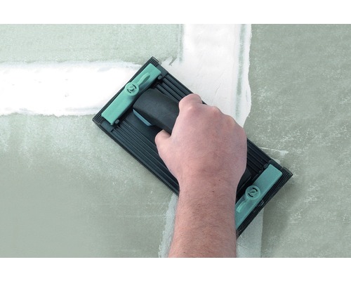 Een hand gebruikt een schuurplank om een geplamuurde muur glad te maken.