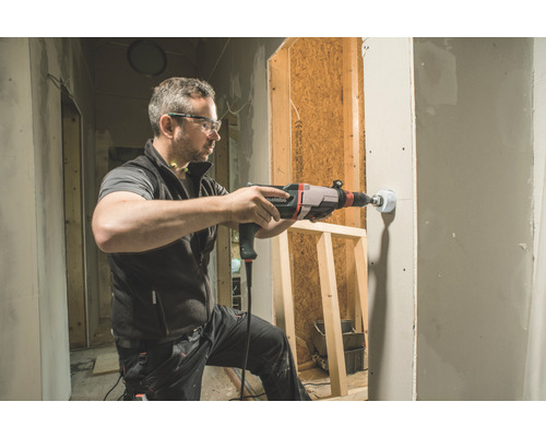 Een man boort met een boormachine een gat in een wand. De scène toont een bouwplaats.